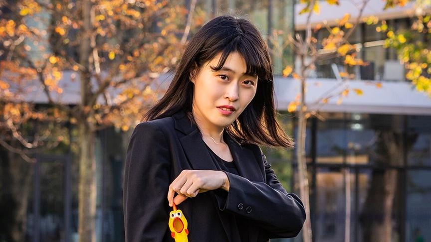 © ORF/Ö1/Kazuki Ikegami Reina Yoshioka steht im Freien vor einem modernen Gebäude und hält eine gelbe Gummihenne vor der Tasche ihres schwarzen Blazers.