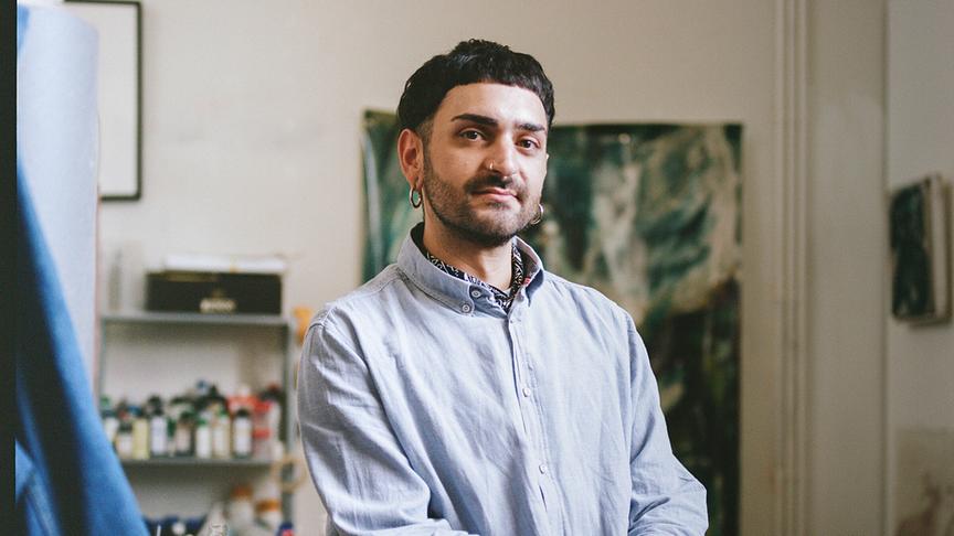 Christian Azzouni mit Piercings sitzt in seinem Atelier auf einem Tisch und blickt in die Kamera.