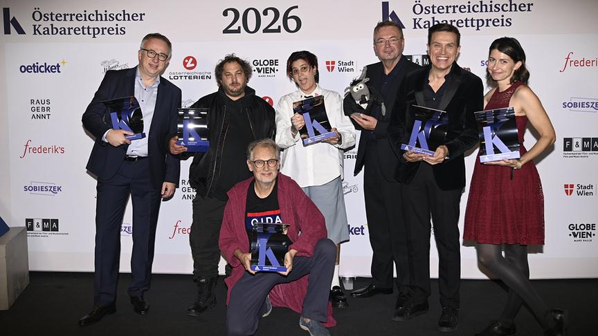 Peter Klien, David Scheid, Schwester Ebra, Stefan Gaugusch (alias Rolf Rüdiger), Robert Steiner, Chrissi Buchmasser und Severin Groebner mit ihren Auszeichnungen posieren vor der Sponsorenwand zum Österreichischen Kabarettpreis 2026: Fernsehpreis geht an „Gute Nacht Österreich“