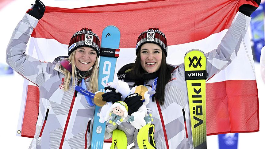 © APA/HANS KLAUS TECHT Die zwei österreichischen Skifahrerinnen Ariane Rädler und Katharina Huber stehen jubelnd mit Goldmedaillen, Skiern und Maskottchen vor einer ausgebreiteten rot‑weiß‑roten Flagge im Rahmen der Siegerehrung der Frauen-Teamkombination in Cortina d'Ampezzo.