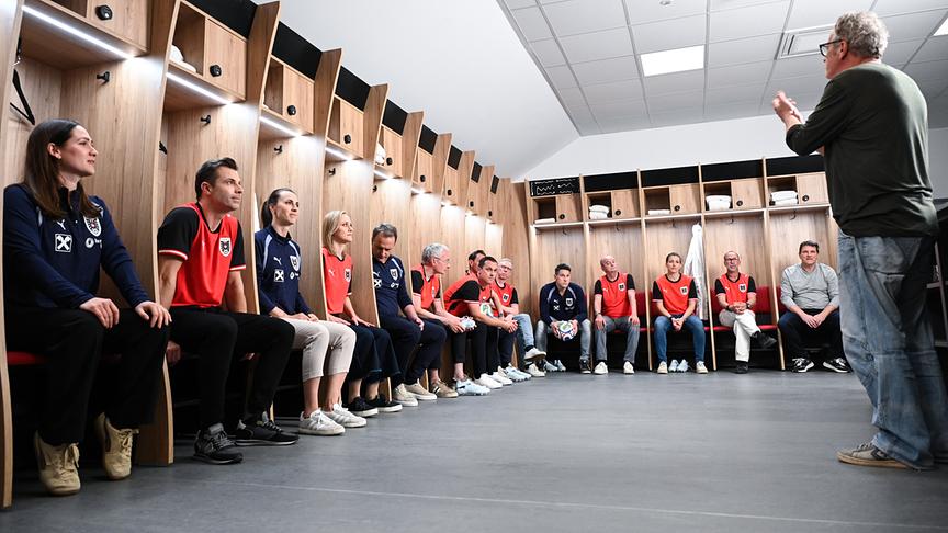 Das ORF-WM-Team sitzt  in Sportkleidung beim Promodreh in der ÖFB-Kabine und hört einem stehenden Mann im Vordergrund zu.