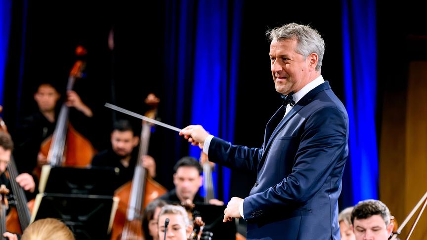 Chefdirigent Markus Poschner  im dunklen Anzug leitet mit seinem Taktstock das ORF Radio-Symphonieorchester Wien auf einer Bühne mit blauem Vorhang. 