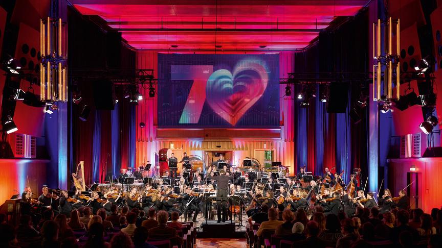 Das Bild zeigt ein großes Orchester, das im farbig ausgeleuchteten Konzertsaal im ORF RadioKulturhaus vor Publikum spielt, während über der Bühne ein leuchtendes 70er-Herzmotiv projiziert wird. 