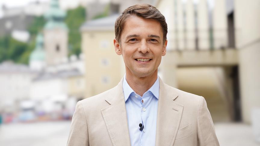 Peter Schneeberger im hellen Sakko und blauen Hemd steht mit Ansteckmikrofon vor der unscharfen Stadtkulisse Salzburgs.
