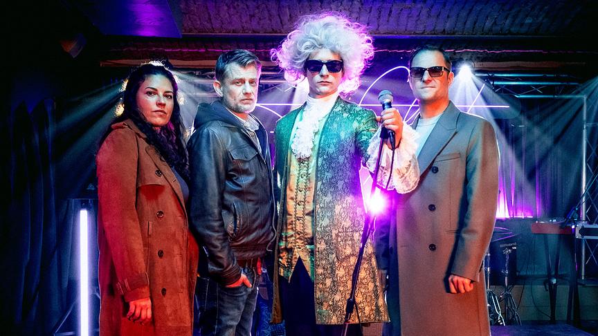 Claudia Kottal, Andreas Guenther, Benedikt Kalcher als Falco im barocken Kostüm mit Mikrofon und Philipp Hochmair stehen auf einer bunt beleuchteten Bühne.