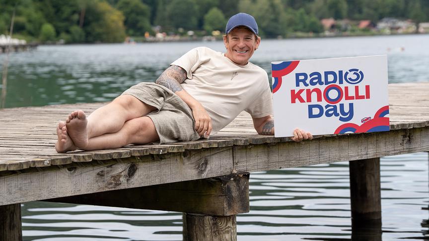 © ORF/Hitradio Ö3/Martin Domkar Ö3-Moderator Andi Knoll liegt auf einem Badesteg am See und präsentiert ein Schild für den „Radio Ö3 Knolliday“.