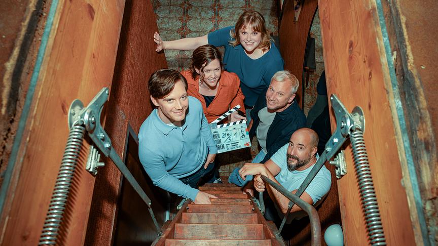 © ORF/Rundfilm/Nikolett Kustos Jonas Nay, Regisseur Christopher Schier, Emily Cox und Robert Stadlober schauen in einem engen, holzverkleideten Dachbodenaufgang nach oben in die Kamera, eine Person hält dabei eine Filmklappe.