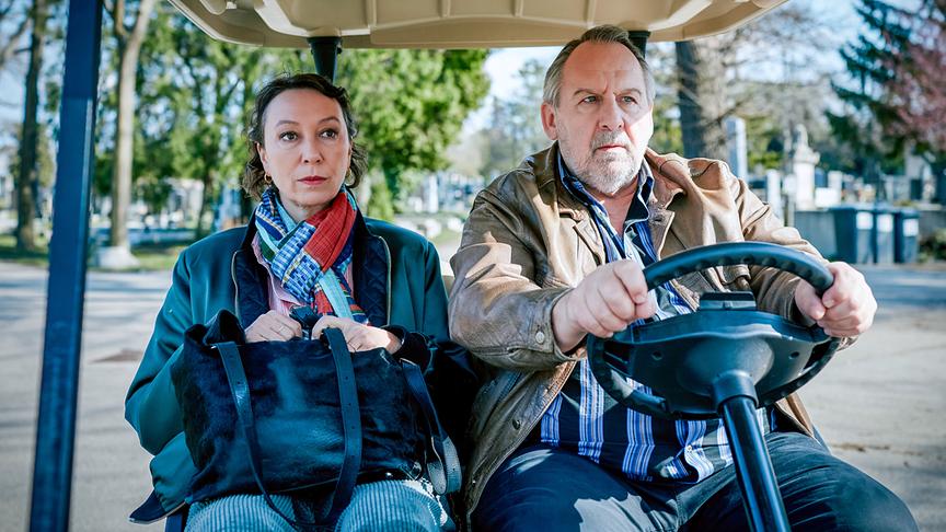 Angelika Schnell und Harald Franitschek sitzen nebeneinander in einem kleinen Elektrofahrzeug und fahren über einen Friedhof.