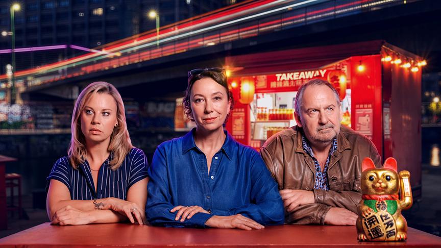 Maja Landauer, Angelika Schnell und Harald Franitschek sitzen vor einem nächtlichen asiatischen Take-away-Lokal an einem Tisch, auf dem eine goldene Winkekatze steht.