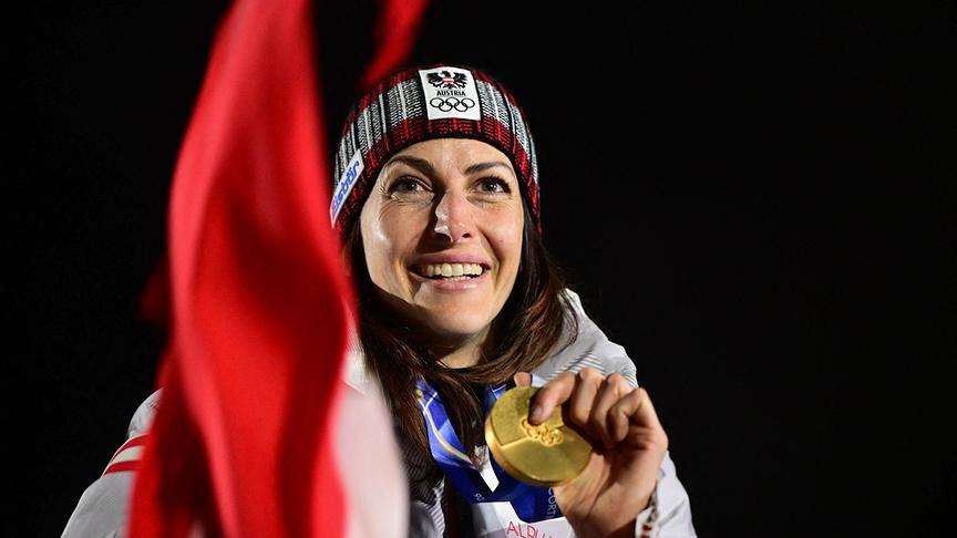 Die österreichische Olympia-Goldmedaillengewinnerin Janine Flock präsentiert lächelnd ihre Medaille auf dem Podium nach ihrem Wettkampf im Skeleton der Frauen, während im Vordergrund unscharf eine rot-weiße Fahne zu sehen ist.