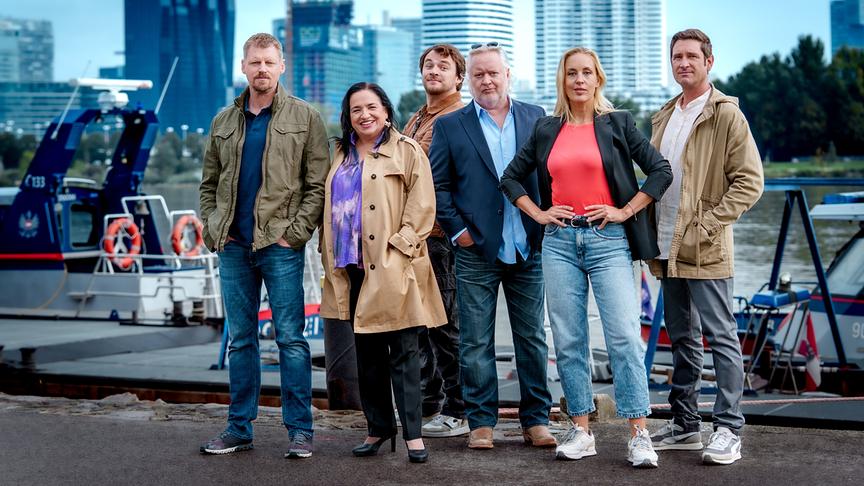 Martin Gruber, Maria Happel, Max Fischnaller, Gregor Seberg, Lilian Klebow und Andreas Kiendl stehen am Ufer vor einem Boot, im Hintergrund ist die Skyline von Wien zu sehen.