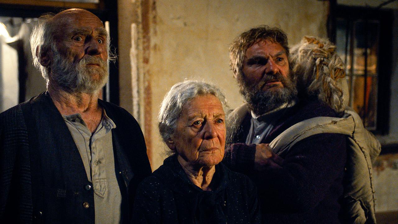 Isidor Falkner, Adelheid Falkner und Romed Falkner mit einer Stoffpuppe stehen nebeneinander in einem alten Bauernhaus und schauen ernst in eine Richtung.