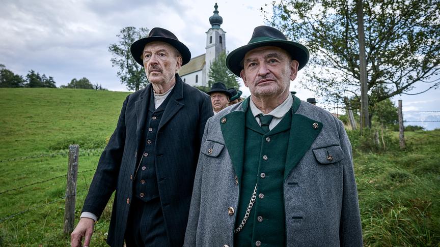 © ORF/ZDF/Claussen+Putz/Film AG/Fabio Eppensteiner Josef Hader und Sigi Zimmerschied schauen gerade aus, im Hintergrund eine Dorfkirche auf einer Anhöhe