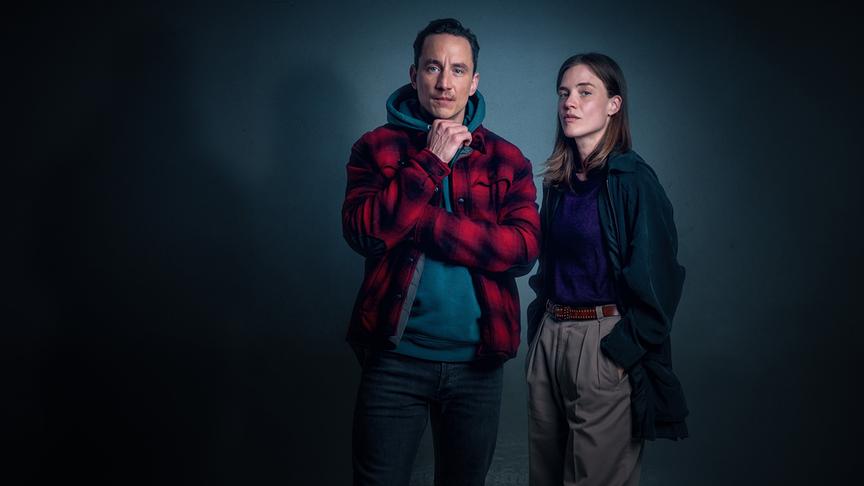 Miriam Fussenegger und Laurence Rupp als neues "Tatort"-Ermittlungsduo Charlie Hahn und Alex Maleky stehen in lässiger Kleidung vor einem dunkelblauen Hintergrund und blicken ernst in die Kamera.