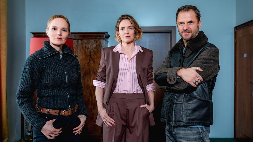 © ORF/ZDF/Graf Film/Petro Domenigg Gruppenfoto mit Julia Koch, Patricia Aulitzky und Philipp Hochmair in einem schlichten Raum mit blauer Wand.