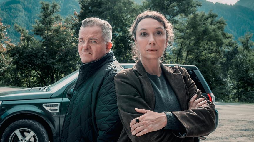 Ursula Strauss als Fanny Gruber und Cornelius Obonya als Benedikt Renner stehen vor einem grünen Geländewagen und blicken direkt in die Kamera, im Hintergrund Bäume und bewaldete Berge