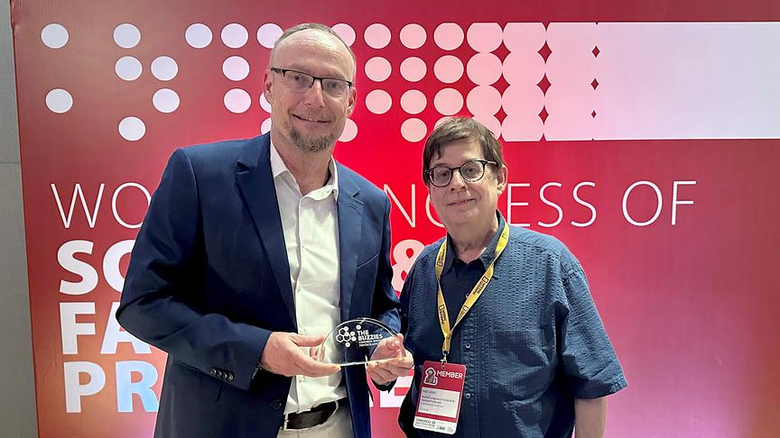 © ORF Genrot Lercher und Paul Lewis stehen vor einer rot gestalteten Kongresswand, wobei eine von ihnen eine gläserne Auszeichnung mit der Aufschrift „The Buzzies“ präsentiert.