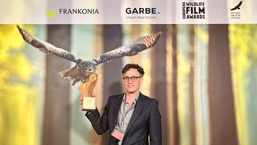© ORF Universum-Regisseur und Kameramann Patrick Centurioni hält vor einer Fotowand den Preis für den „Besten Europäischen Film“ in der Kategorie „Conservation/Naturschutz“. in die Höhe; oben sind Logos wie „European Wildlife Film Awards“ zu sehen und im Hintergrund ist eine große Eule mit ausgebreiteten Flügeln abgebildet.