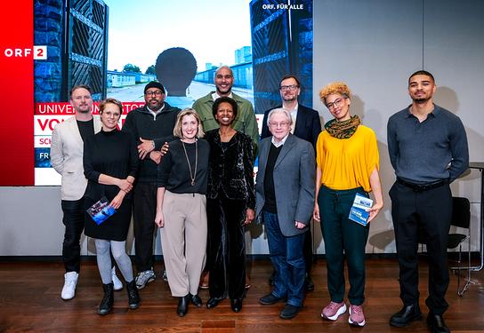 © ORF/Roman Zach-Kiesling Caroline Haidacher, Stefanie Daubek, Mary L. Romney-Schaab, Walter Sauer, Claudia Unterweger, Jeremias Amoore, Clemens Radauer, David Mayonga, Jermain Raffington und Gerald Heidegger posieren bei der „Universum History“-Neuproduktion „Vom Paradies in die Hölle – Schwarze Menschen im Nationalsozialismus“ in einem ORF-Studio vor einer großen Bildwand mit ORF2‑Grafik und Sendungstitel.