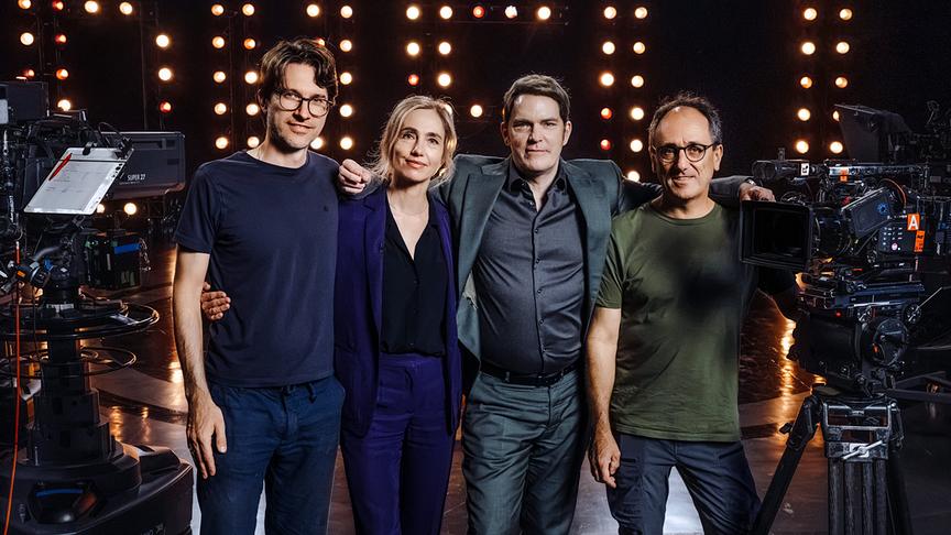 Regisseur Rudi Gaul, Ursina Lardi, Godehard Giese und Kameramann Stefan Sommer stehen zwischen Studiokameras vor einer Wand aus warm leuchtenden Scheinwerfern bei den Dreharbeiten zum „Duell“ (AT).