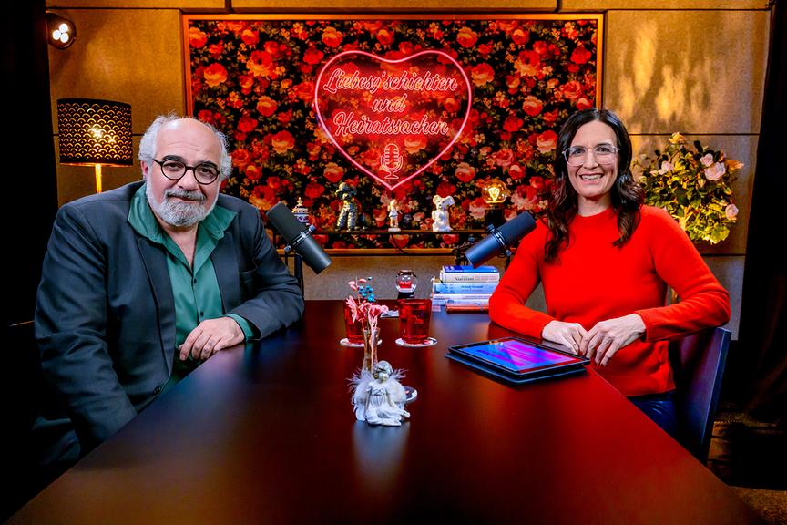 Michael Niavarani und Nina Horowitz sitzen an einem dunklen Holztisch beim ORF-„Liebesg’schichten und Heiratssachen – Der Podcast“ mit Nina Horowitz
