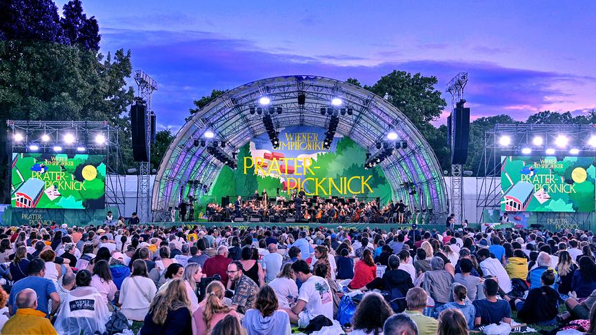 © ORF/Klaus Titzer Eine große Menschenmenge sitzt bei Abenddämmerung vor einer beleuchteten Open-Air-Bühne, auf der die Wiener Symphoniker beim „Prater‑Picknick“ unter freiem Himmel auftreten.