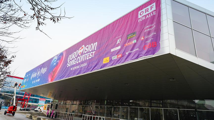 Ansicht der Wiener Stadthalle, wo das Vordach mit einem großen Banner zum „Eurovision Song Contest – Vienna 2026“ mit ORF-Branding behängt ist.