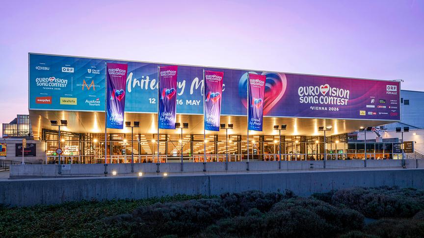 Abendlich beleuchtete Wiener Stadthalle mit großen Bannern zum Eurovision Song Contest Vienna 2026 vor violettem Abendhimmel.