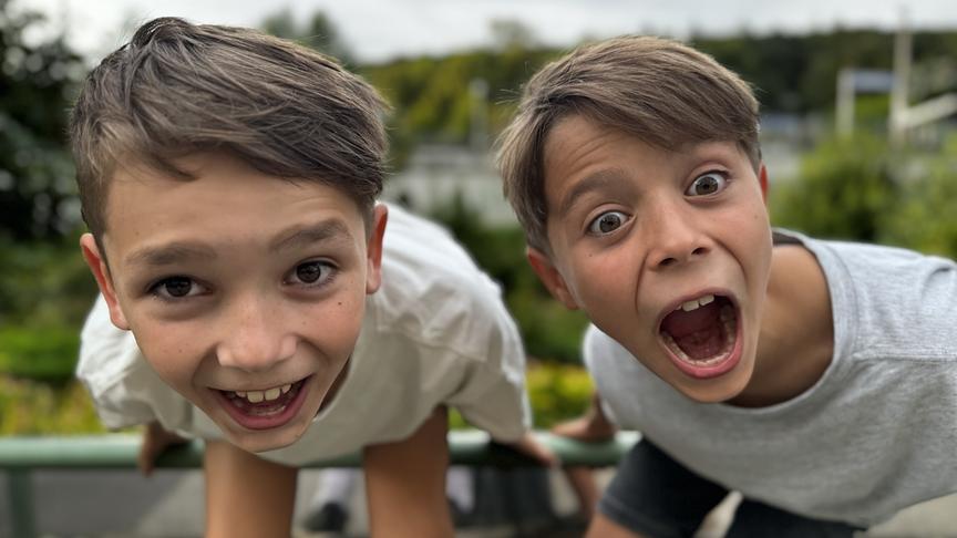 "Die Abenteuerchecker" Benni und Gucki beugen sich im Freien über ein Geländer, während im Hintergrund eine grüne, leicht unscharfe Landschaft zu sehen ist.