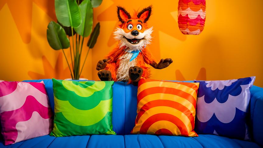 © ORF/Steiner Familyentertainment/Florian Falb Fuchsi, eine fröhliche Fuchs‑Handpuppe, lehnt an einem Tisch in einem bunt gestalteten Studio mit Regalen und Pflanzen im Hintergrund.