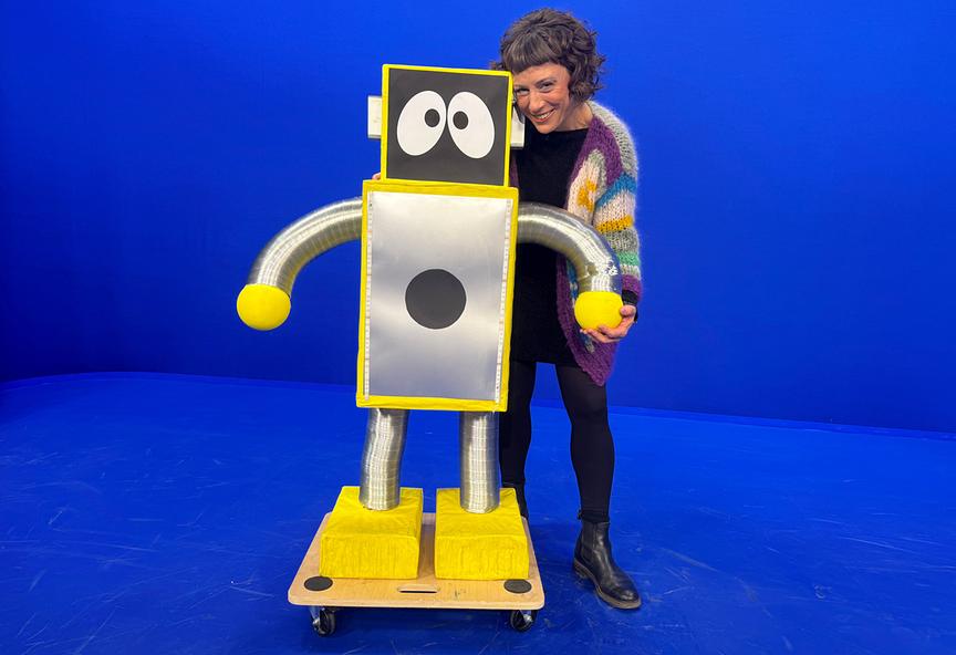 Esther und KIKI, eine selbstgebaute, gelb‑silberne Roboterfigur mit großen Augen, stehen in einem Studio mit blauem Hintergrund.