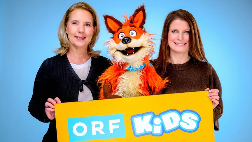 Alexandra Schlögl und Yvonne Lacina-Blaha halten gemeinsam mit Fuchsi, fröhlichen Fuchs‑Handpuppe, ein gelbes Schild mit dem Logo „ORF KiDS“ vor blauem Hintergrund.