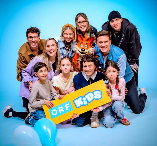 Christoph Hirschler, Melanie Flicker, Fanny Stapf, Lena Großmann, Esther Csapo, Robert Steiner, Stefan Gruber mit Fuchsi und Kindern posieren vor blauem Hintergrund und halten ein Schild mit der Aufschrift „ORF Kids“.