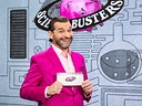 Martin Puntigam im pinken Anzug steht vor dem „Science Busters“-Logo und hält eine Moderationskarte.
