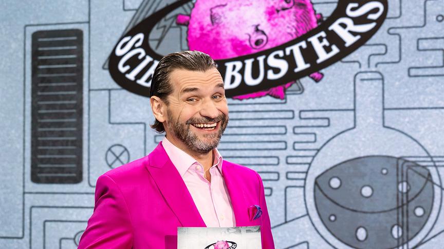 Martin Puntigam im pinken Anzug steht vor dem „Science Busters“-Logo und hält eine Moderationskarte.