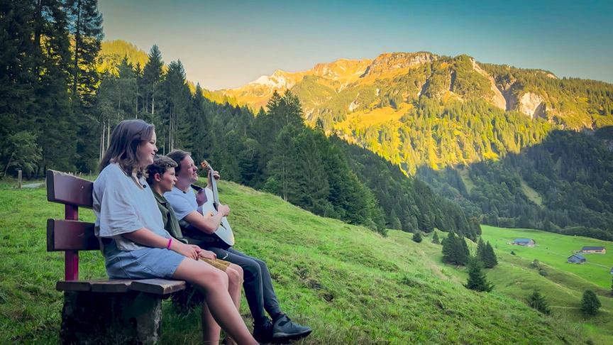 Musiker Philipp Lingg singt mit Nichte Heidi und Neffen Jonathan ein typisches Lied aus dem Bregenzer Wald auf einer Bank auf einer grünen Almwiese mit Blick auf eine sonnig beleuchtete Berglandschaft mit Wäldern und Hütten.