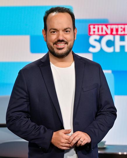 Patrick Budgen in dunkelblauem Sakko, weißem T‑Shirt und Jeans steht im TV‑Studio von "Hinter den Schlagzeilen" vor einem blau gestalteten Hintergrund mit Sendungslogo und moderner Sitzgelegenheit.