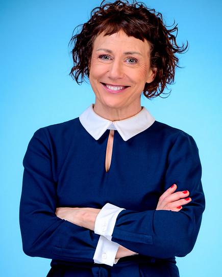 Esther Csapo in blauem Kleid mit weißem Kragen steht vor hellblauem Hintergrund