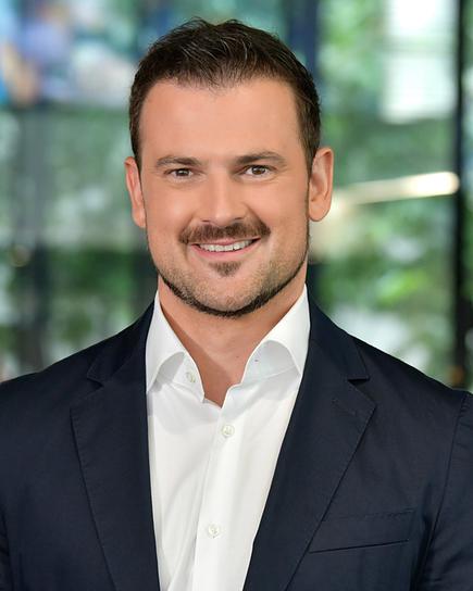 Sigi Fink in dunklem Anzug und weißem Hemd steht im modernen, hellen Newsroom mit unscharfem Hintergrund.