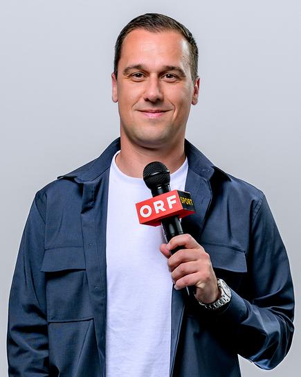 Michael Liendl steht vor grauem Hintergrund und hält ein ORF-Mikrofon in der Hand.