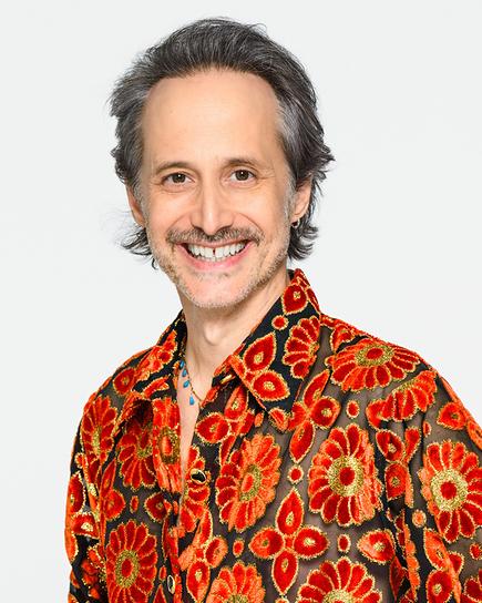 Michael Ostrowski posiert vor weißem Hintergrund in einem schwarz-rot-gold-gemusterten Hemd kombiniert mit schwarzer Lederhose.