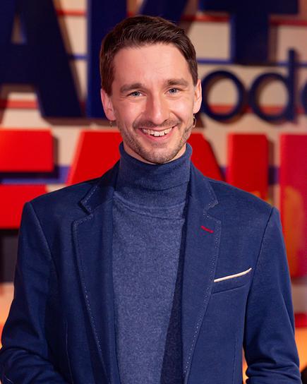 In einem Fernsehstudio steht Moderator Clemens Maria Schreiner lächelnd vor dem Schriftzug „FAKT oder FAKE“.