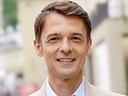 Peter Schneeberger im hellen Sakko und blauen Hemd steht mit Ansteckmikrofon vor der unscharfen Stadtkulisse Salzburgs.