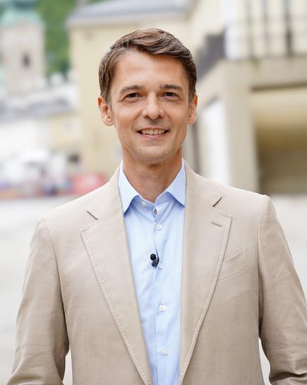 Peter Schneeberger im hellen Sakko und blauen Hemd steht mit Ansteckmikrofon vor der unscharfen Stadtkulisse Salzburgs.