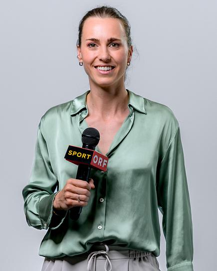 Viktoria Schnaderbeck hält lächelnd vor neutralem Hintergrund ein Mikrofon in die Kamera.