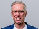 Peter Stöger steht vor grauem Hintergrund und hält ein ORF-Mikrofon in der Hand.