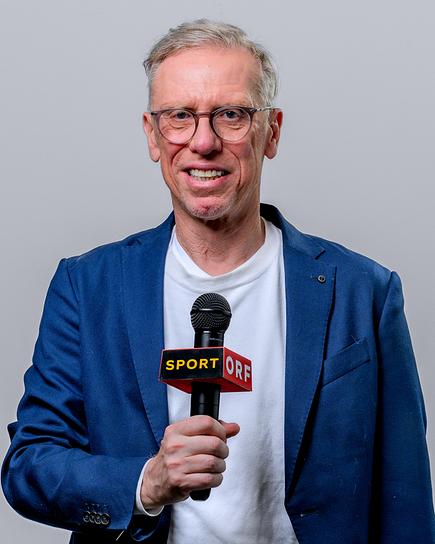 Peter Stöger steht vor grauem Hintergrund und hält ein ORF-Mikrofon in der Hand.